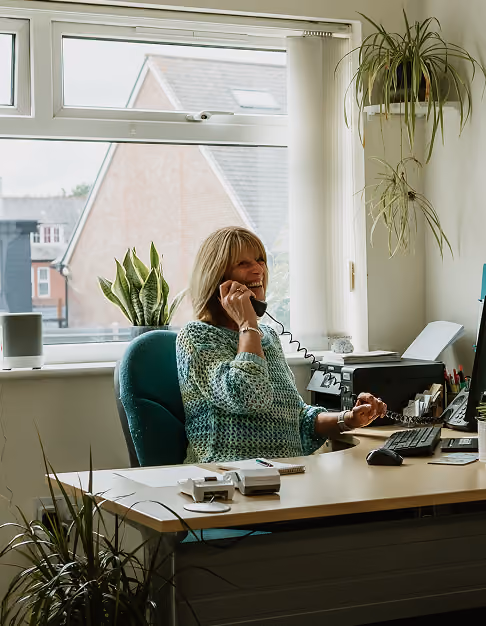 A female receptionist wearing a patterned sweater sits at a desk with a corded phone and computer monitor in a physiotherapist reception area, with a large window and potted plants behind her.