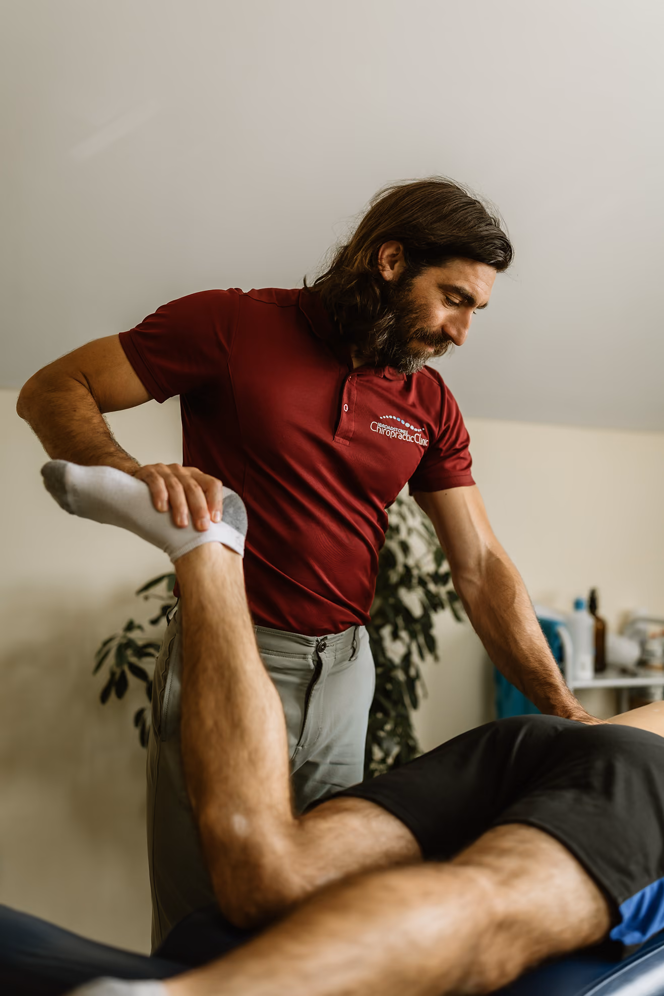 Chiropractor in maroon shirt assisting patient by stretching their leg on a treatment table.