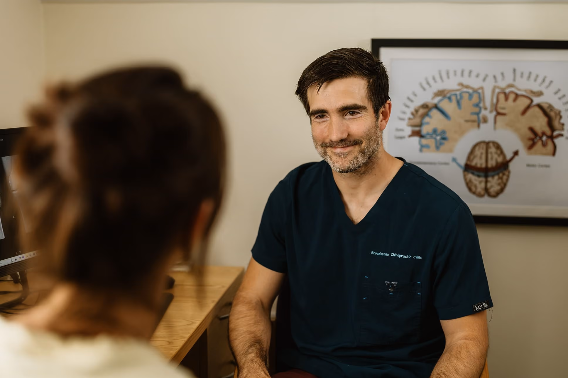 Male physiotherapist in navy scrubs talking to a seated patient in a clinical office.