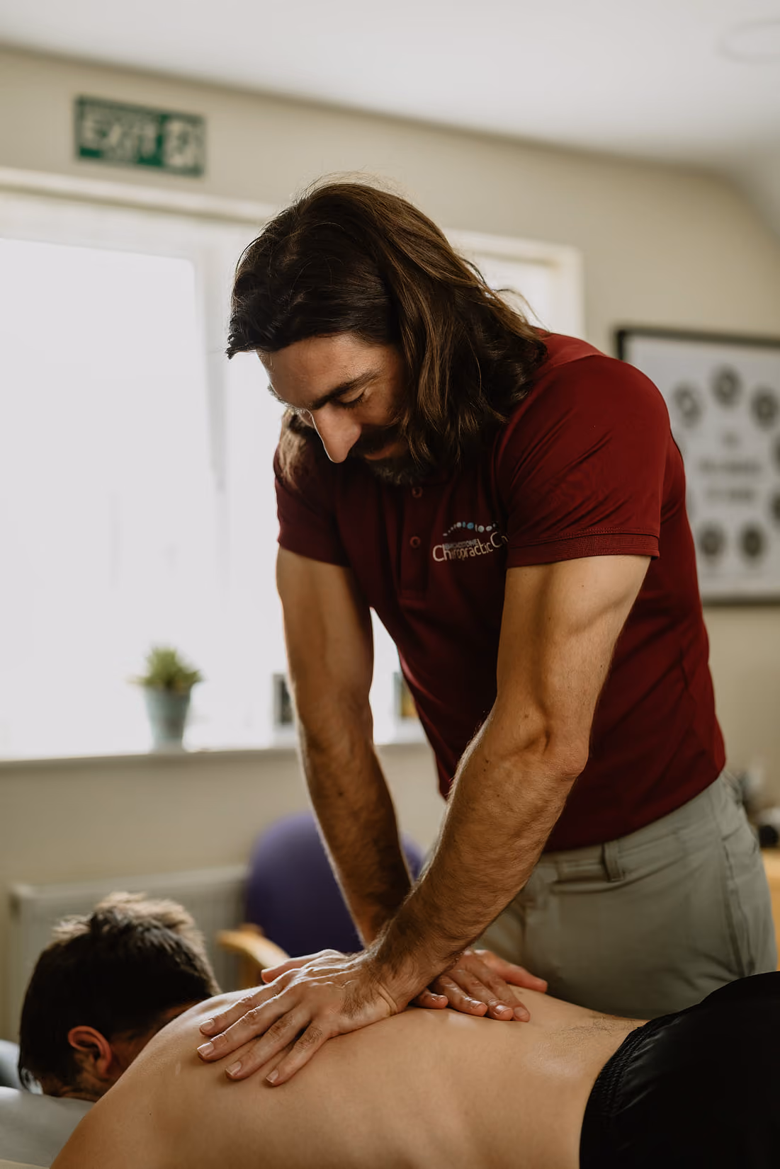 Chiropractor in a maroon shirt giving a back massage to a man lying face down.
