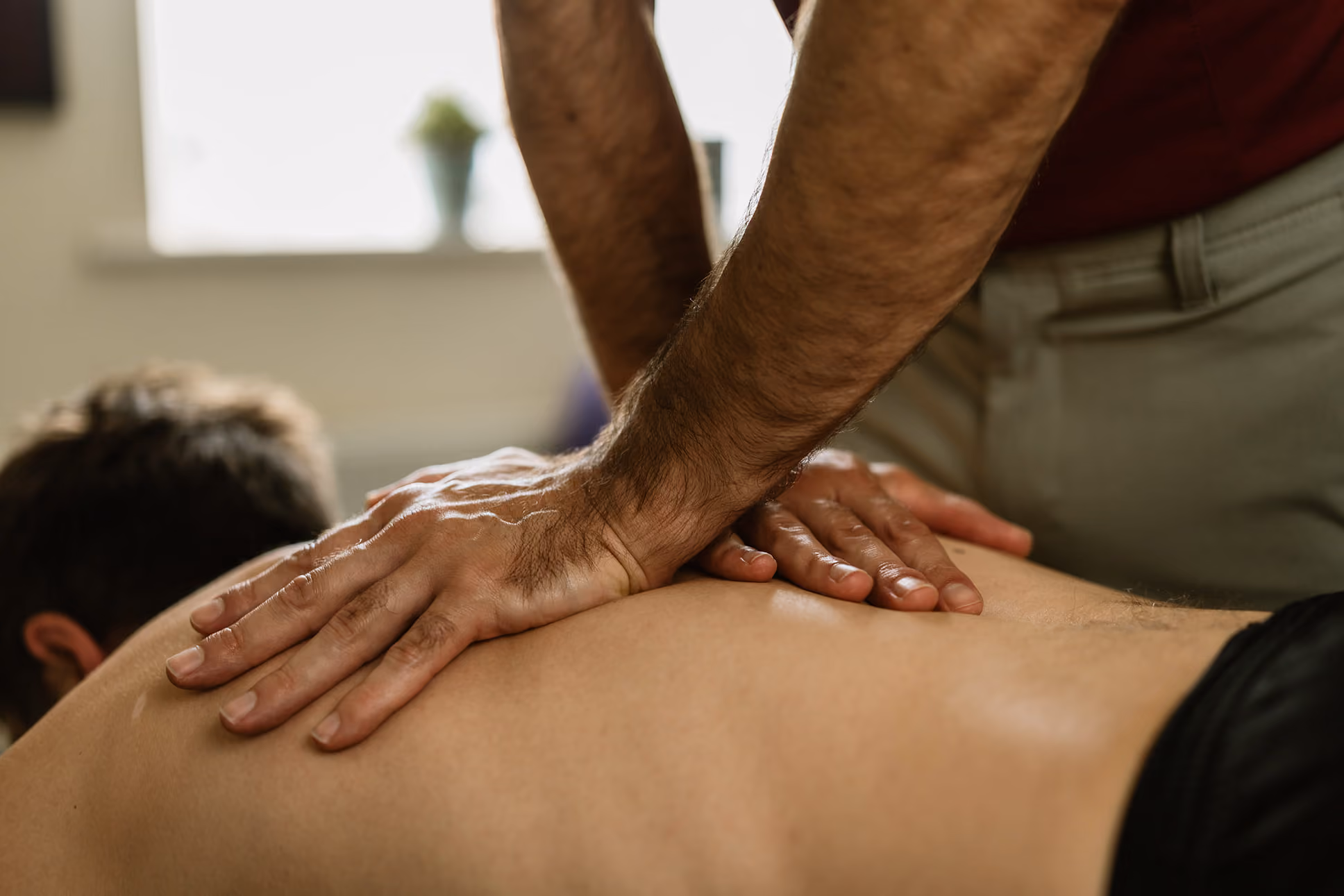 Close-up of hands massaging a man's bare back in a bright room.