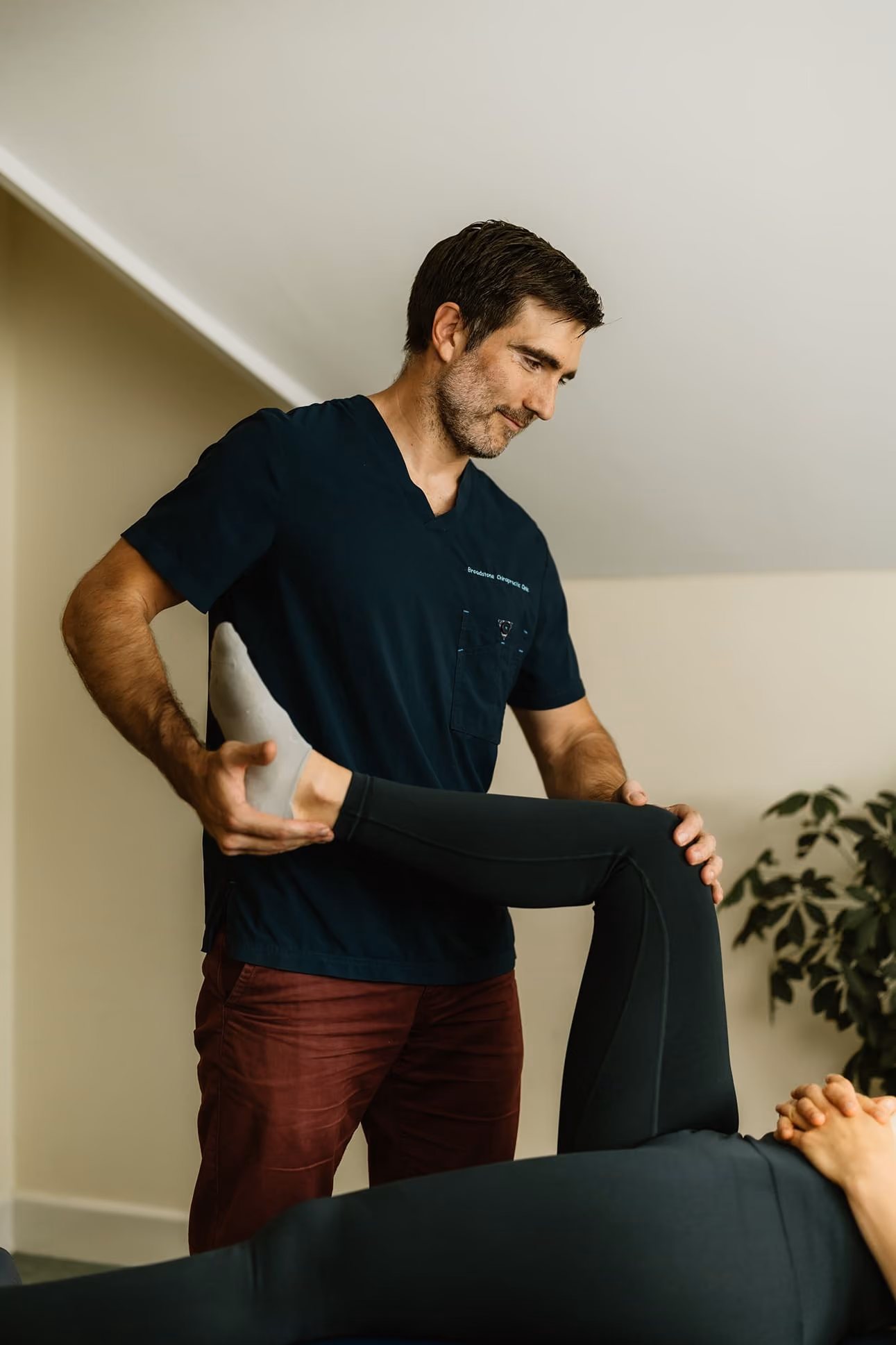 Chiropractor wearing navy scrubs assisting patient by stretching her bent leg in a therapy room.