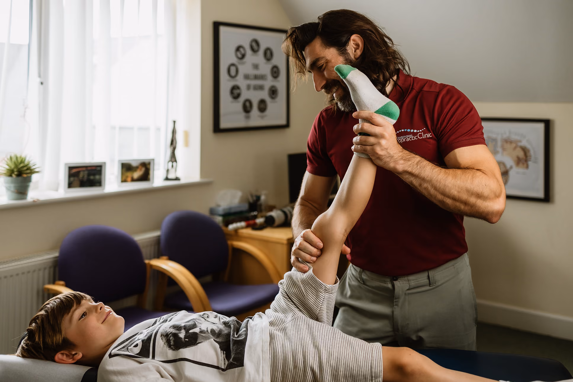 Chiropractor stretching a boy's leg on a treatment table in a clinic with framed medical art on walls.