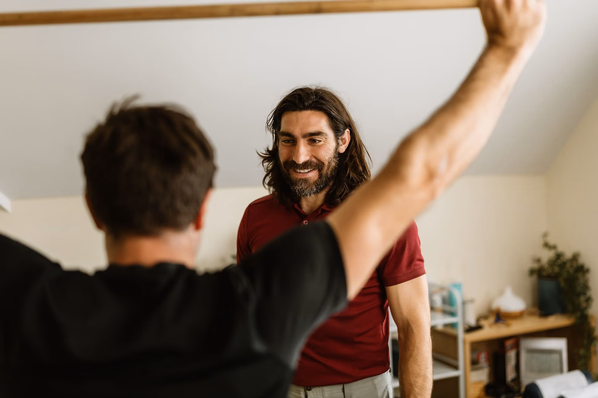 Man in red shirt smiling as another man raises arms in an indoor room with shelves and plants.