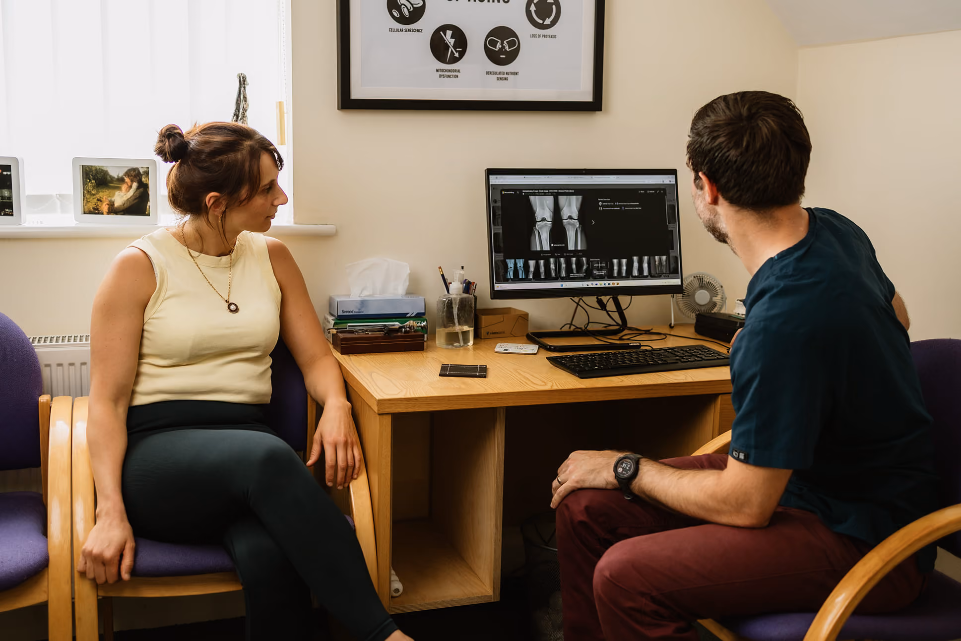 Chiropractor showing a woman X-ray images of knees on a computer screen in a consultation room.
