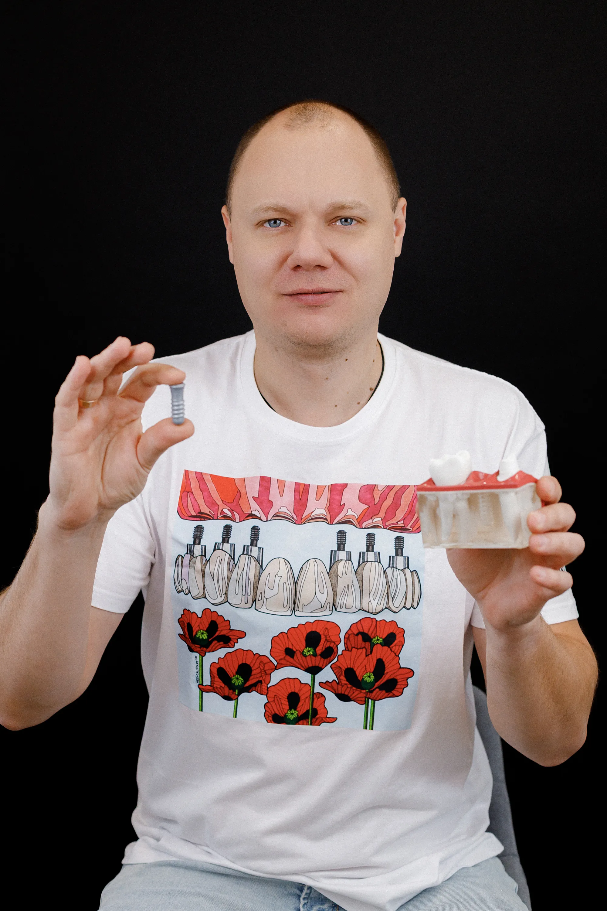 Man holding a dental implant in one hand and a teeth model in the other against black background.