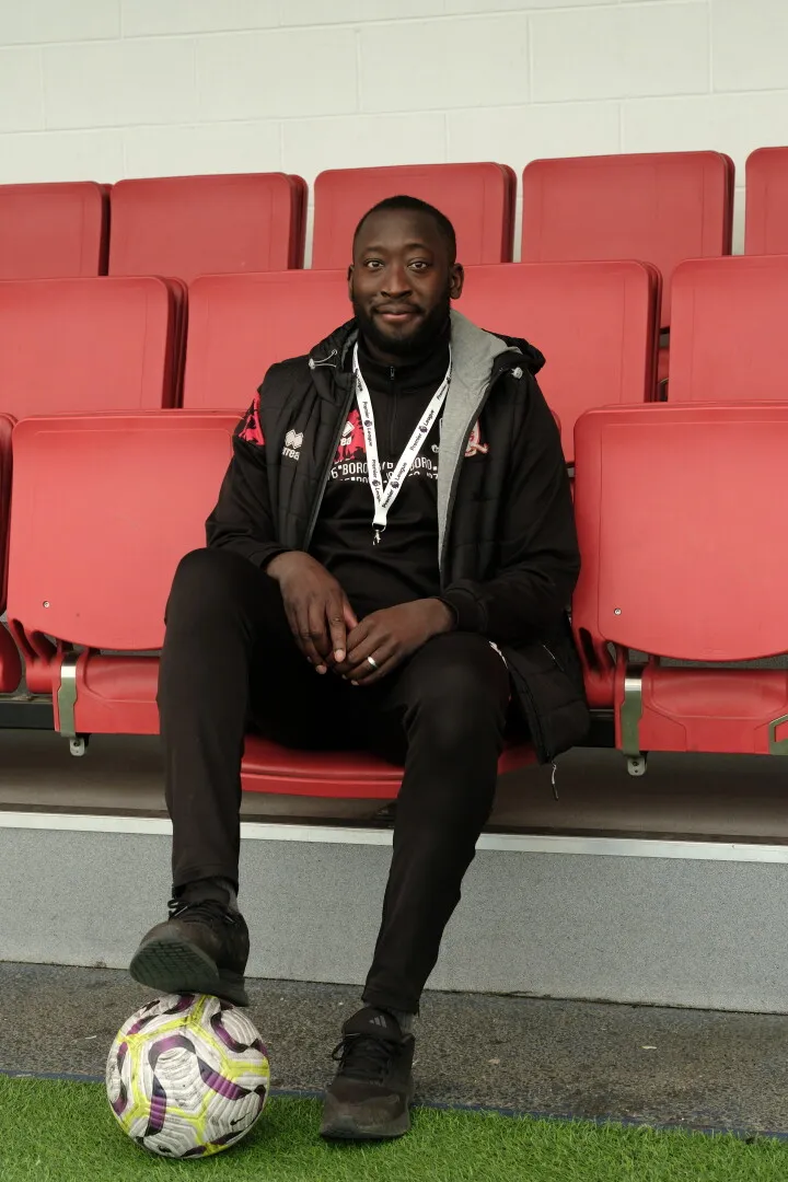Man dressed in black sportswear sitting on a red stadium seat with one foot resting on a football.