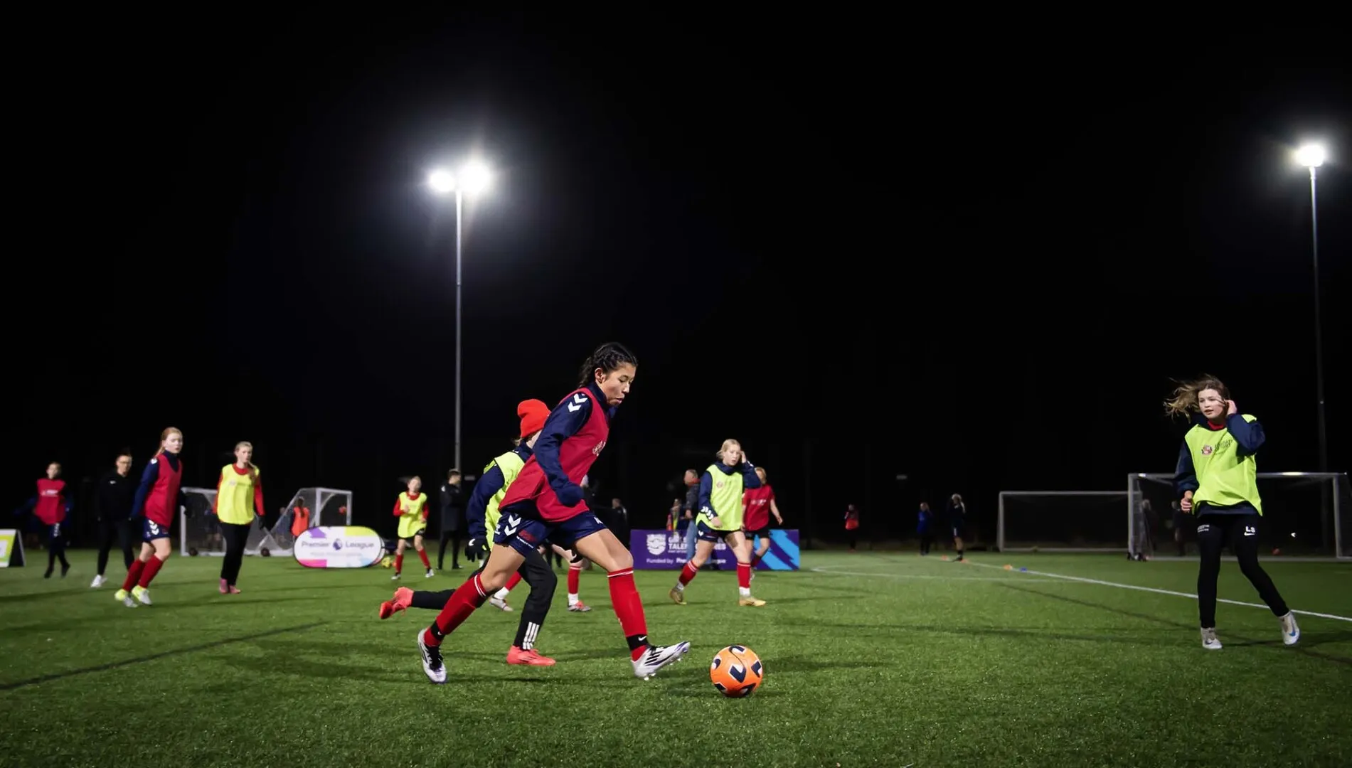 Youth soccer players in red and yellow bibs playing a match under bright floodlights on a green field at night.