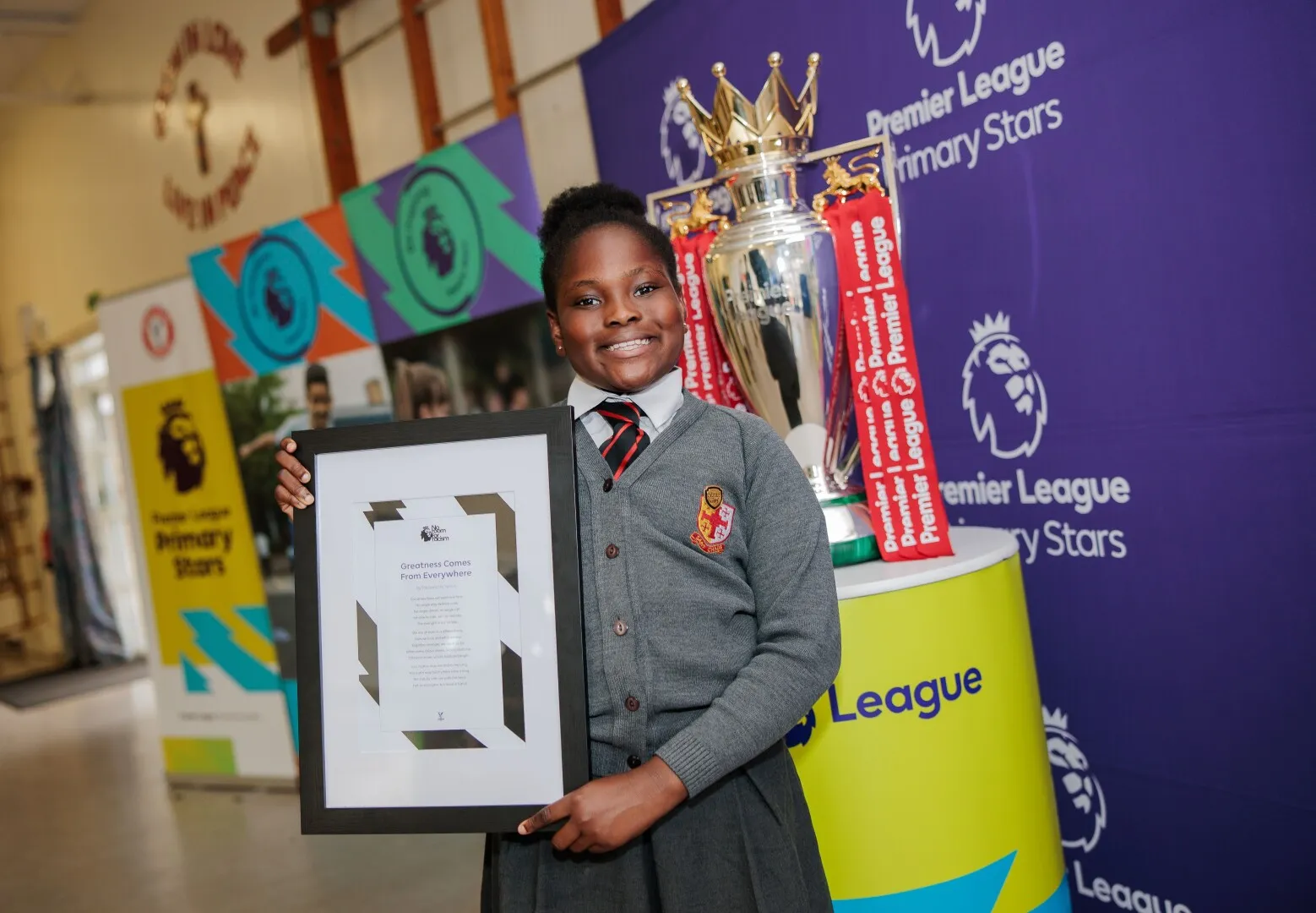 Smiling schoolgirl in uniform holding a framed certificate, standing next to the Premier League trophy at a Primary Stars event.