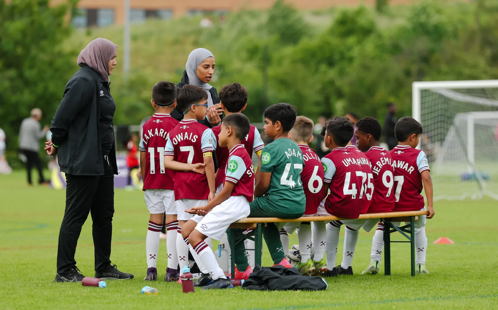 Youth soccer team seated on a bench receiving coaching from two women wearing hijabs on a grassy field near a goalpost.