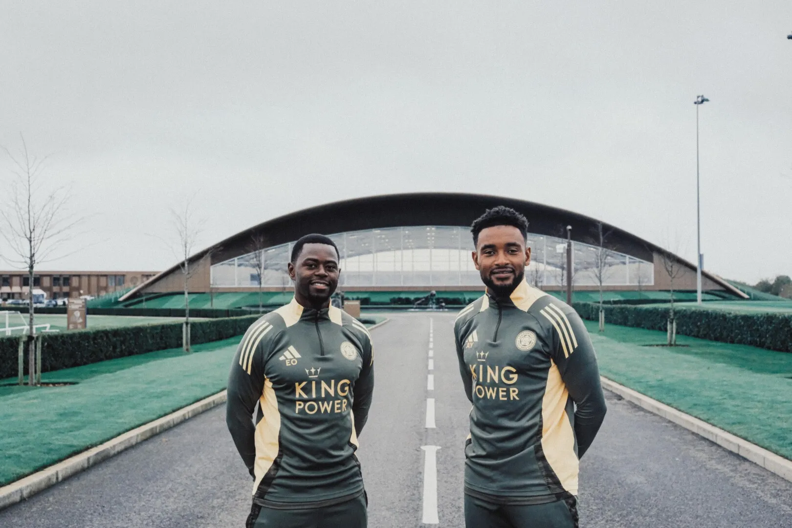 Two men in matching dark athletic jackets with 'KING POWER' on the front standing on a road with a large modern building in the background.