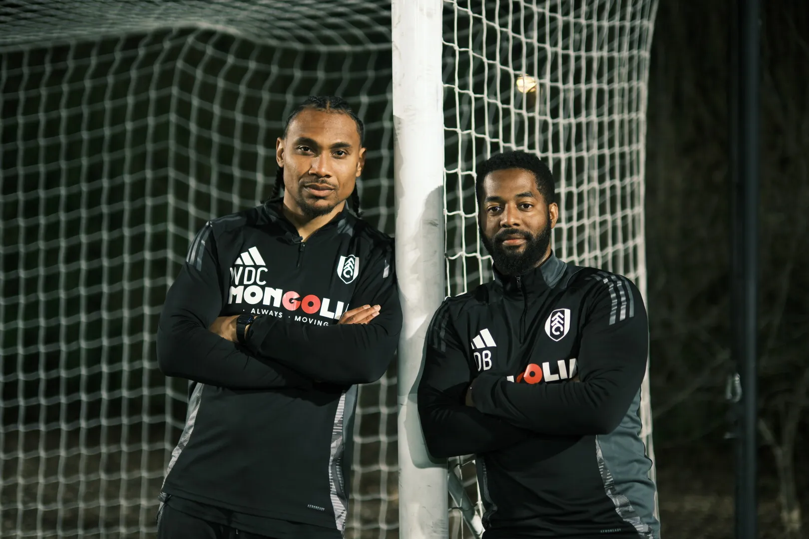 Two men in black athletic jackets with Fulham FC logos leaning against a soccer goalpost at night.