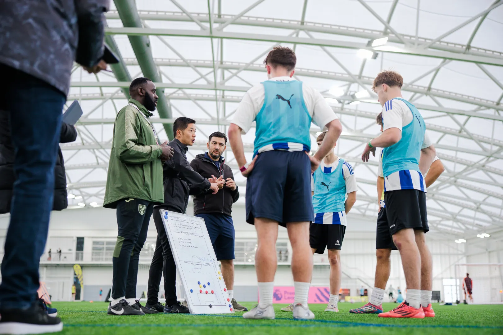 Football coaches and players in blue bibs listening to instructions around a tactics board on an indoor field.