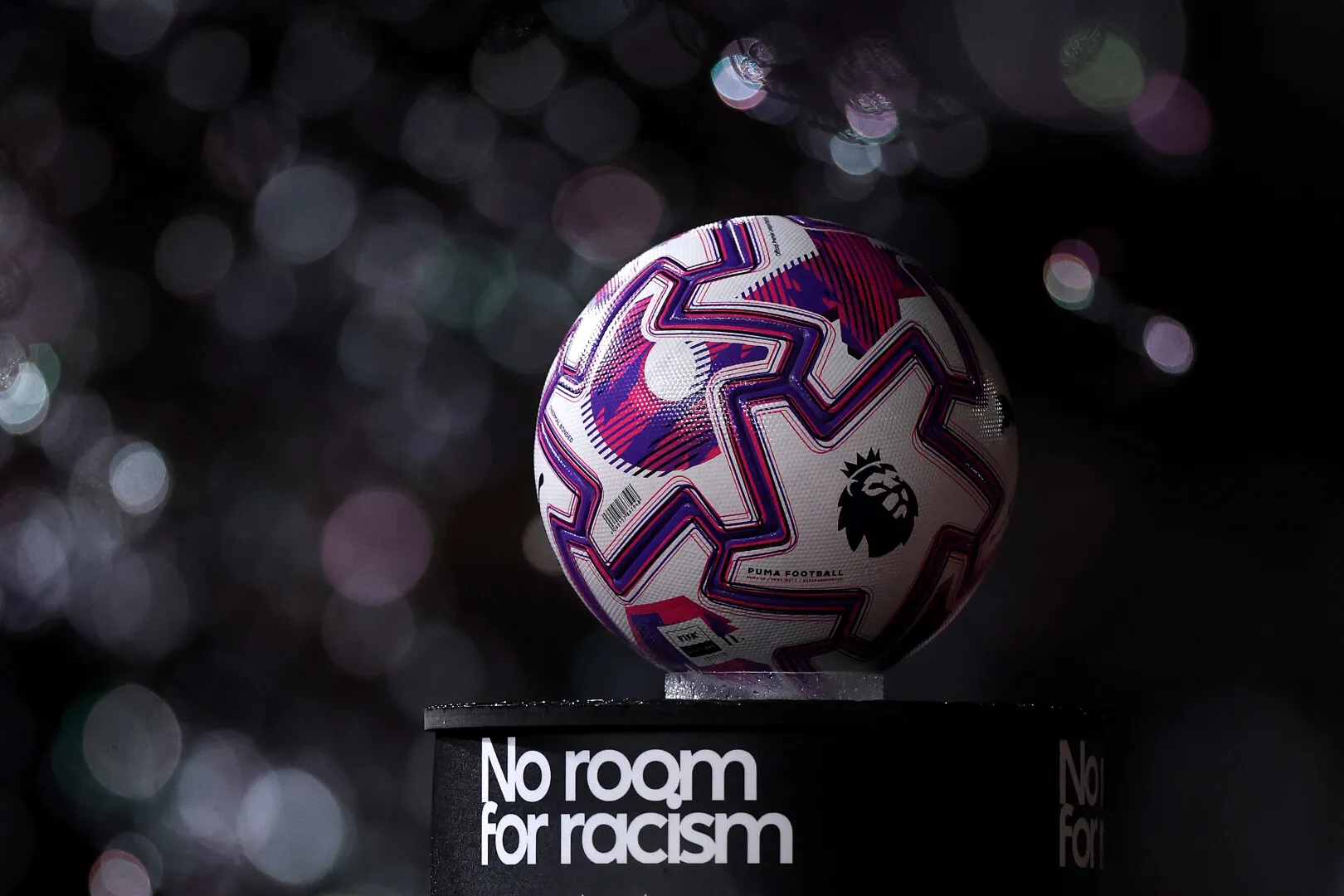 Premier League football with purple and pink patterns on a pedestal labeled 'No room for racism' against a blurred dark background.
