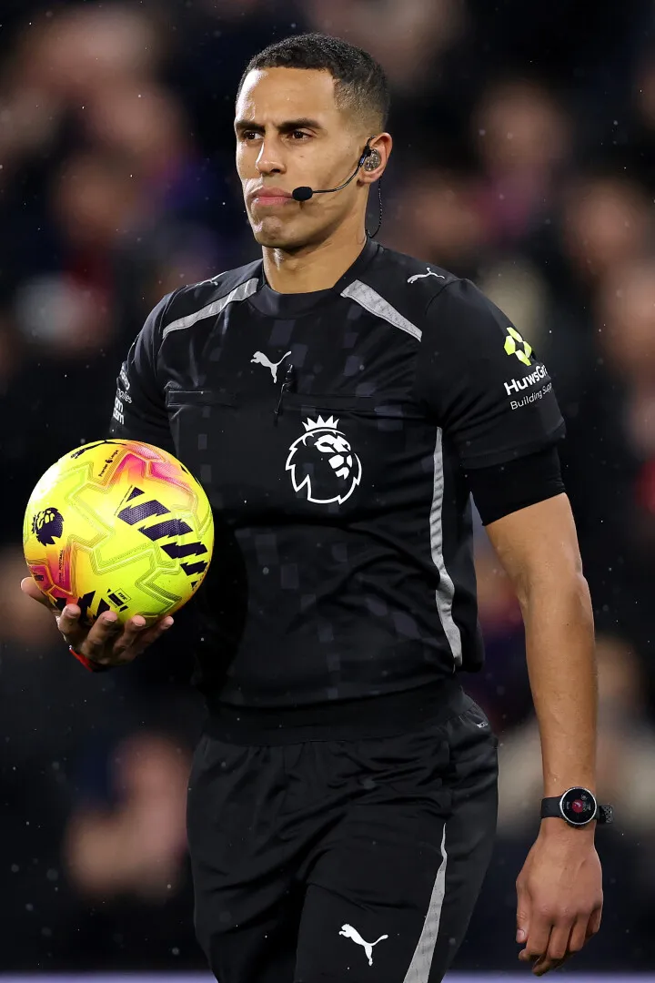 Football referee in black Premier League uniform holding a yellow soccer ball with a focused expression.