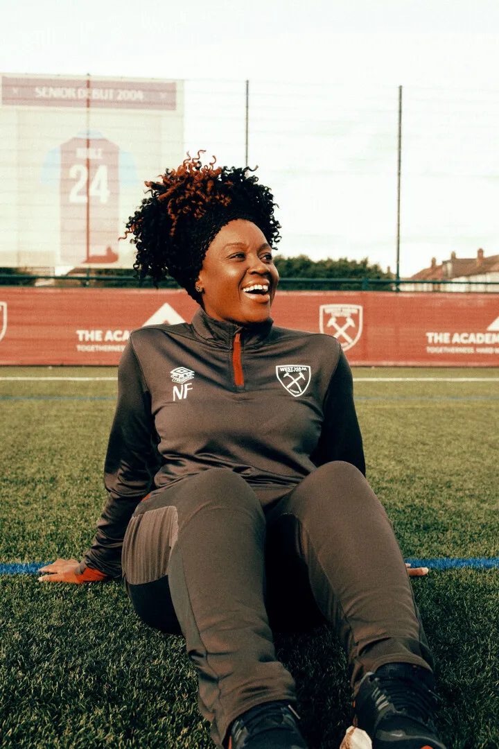Smiling woman in West Ham football training gear sitting on a football field with a blurred banner and fence in the background.