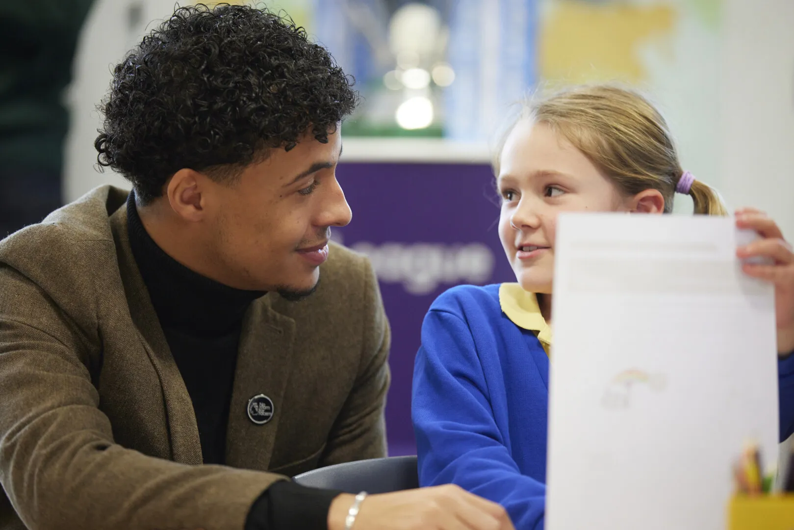 Young man smiling and interacting with a girl in a blue school uniform who is holding up a piece of paper with text and a small rainbow drawing.