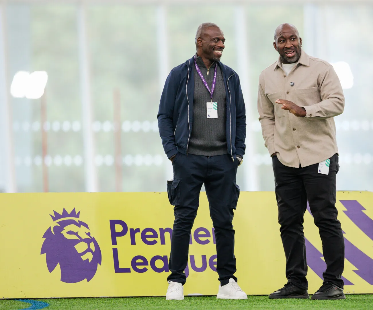 Two men standing on grass next to a yellow Premier League sign with a purple lion logo, engaged in conversation and smiling.