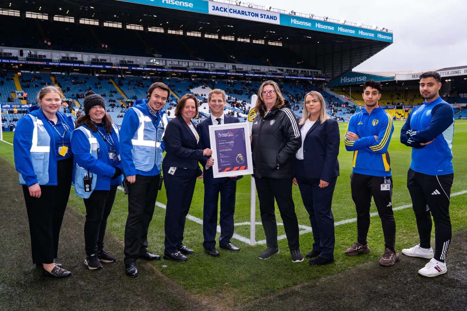 Group of nine people standing on a football pitch at Leeds United's Jack Charlton Stand, holding a framed Premier League certificate.