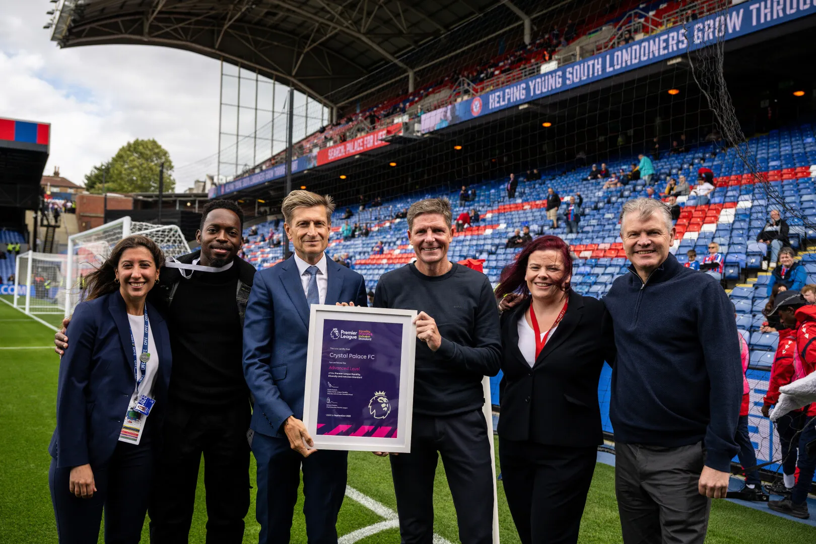 Six people standing on a soccer field with Crystal Palace stadium seats in the background, one holding a framed Premier League Equality award.