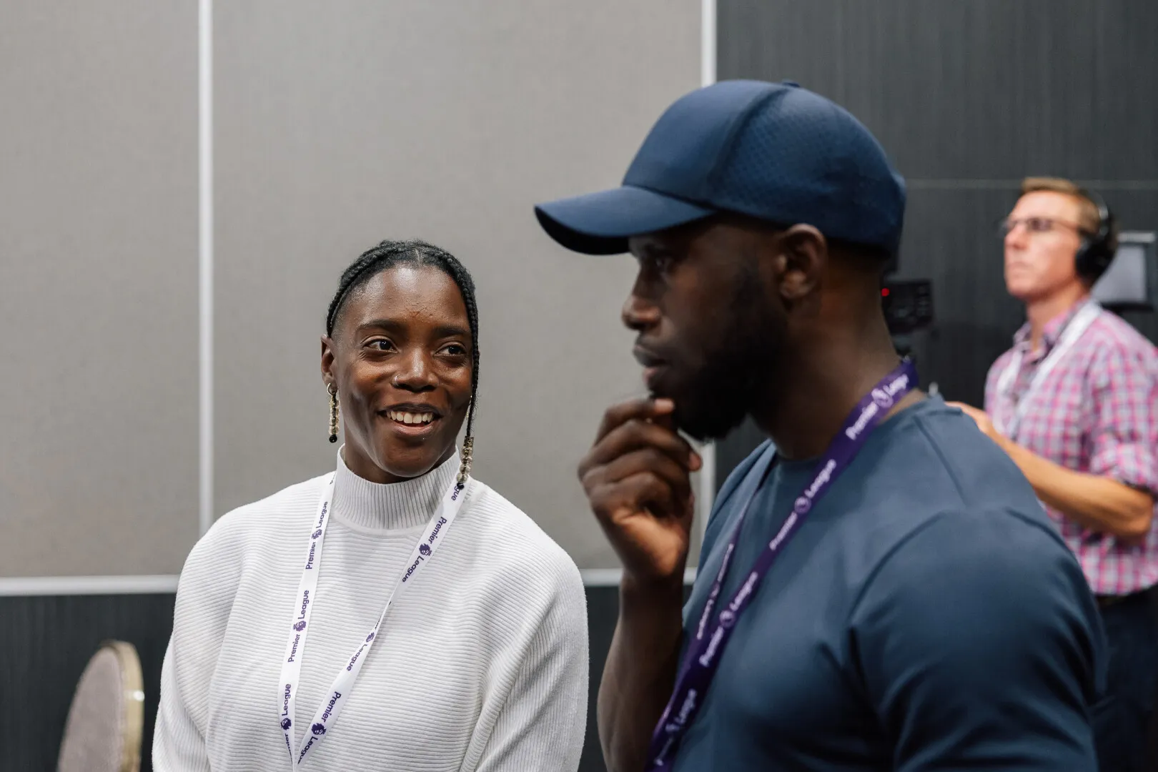 Smiling woman in white sweater and a man in blue shirt and cap wearing Premier League lanyards talking, with a man in checkered shirt and headphones in the background.