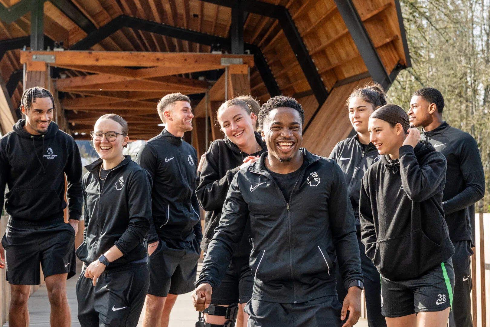 Group of young adults in black sportswear smiling and walking together under a wooden roof structure outdoors.
