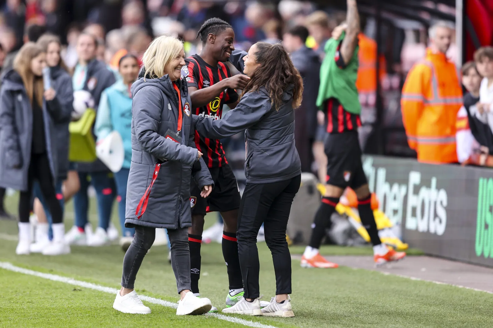 Two women in jackets celebrating with a smiling football player in AFC Bournemouth uniform on a football field.