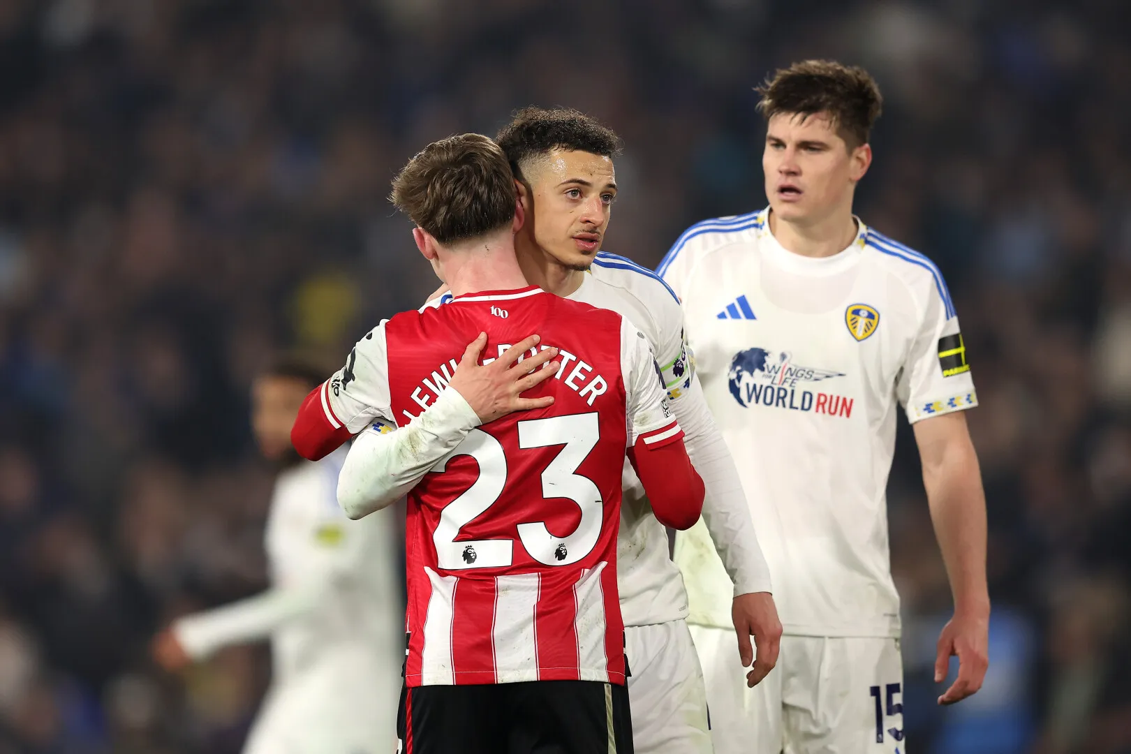Two football players, one in a Sunderland jersey with number 23 and one in a Leicester jersey, embrace on the field while a third player from Leicester watches nearby.