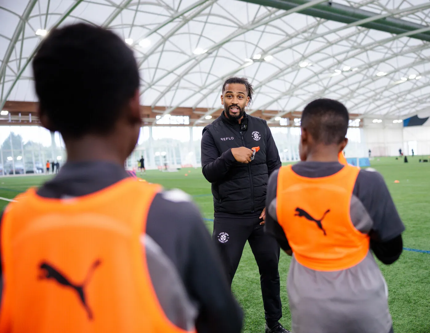 Coach instructing two young soccer players wearing orange training bibs inside an indoor sports facility.
