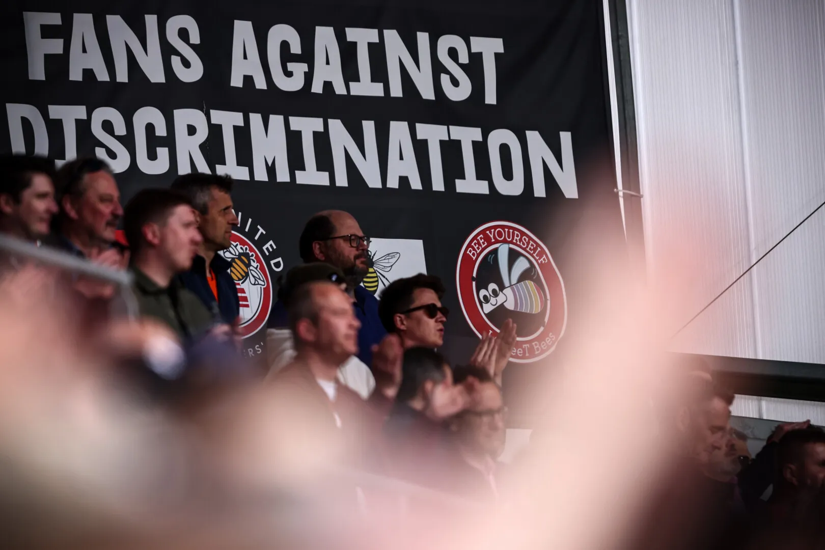 Brentford fans standing and clapping in front of a banner that reads 'FANS AGAINST DISCRIMINATION' next to club logo.