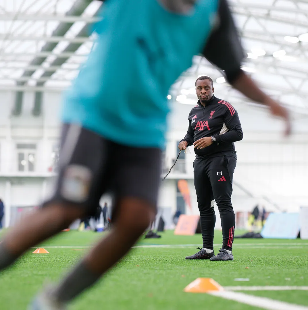 Coach in black Liverpool tracksuit observing a player running during indoor training session on green turf.