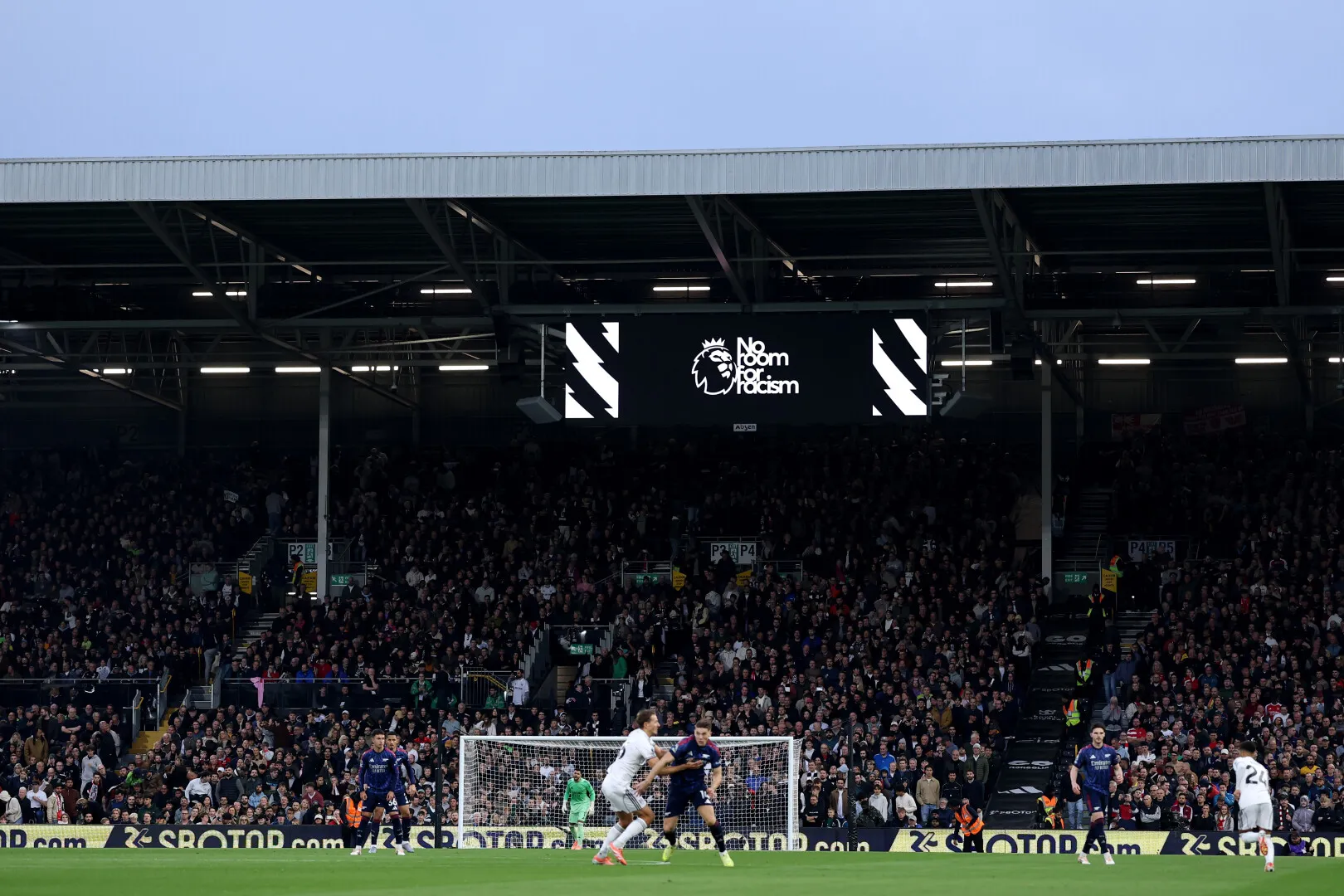 Soccer players competing for the ball in front of the goal with a stadium crowd and a scoreboard displaying 'No room for racism'.
