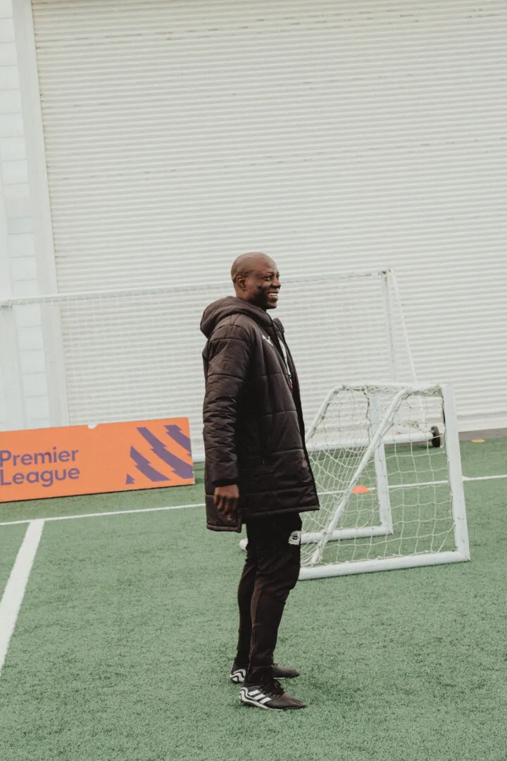 Man in black sportswear and a long coat smiling on a football field near a small goal with a Premier League sign in the background.