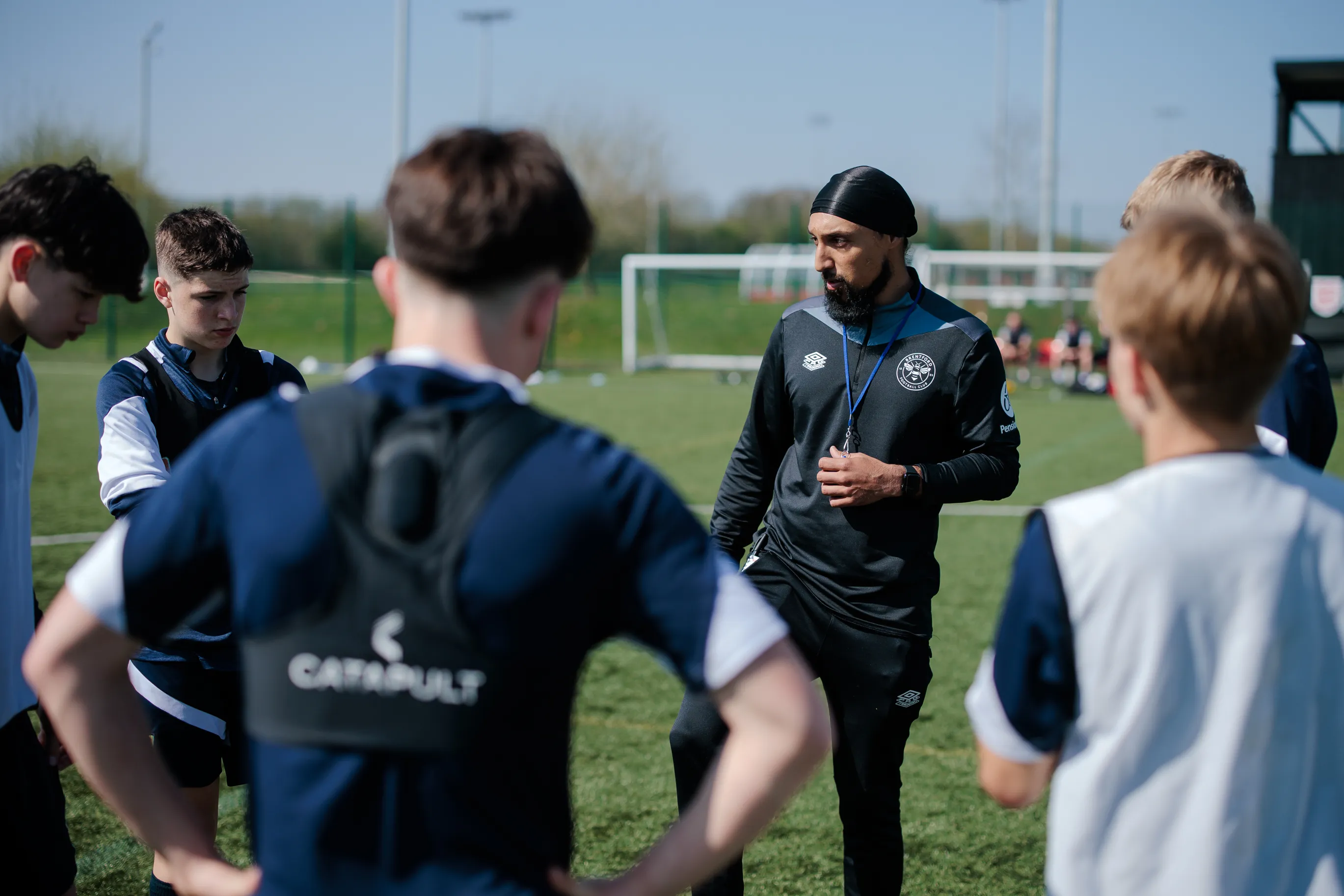 Brentford football coach giving instructions to a group of teenage players on a sunny outdoor field.