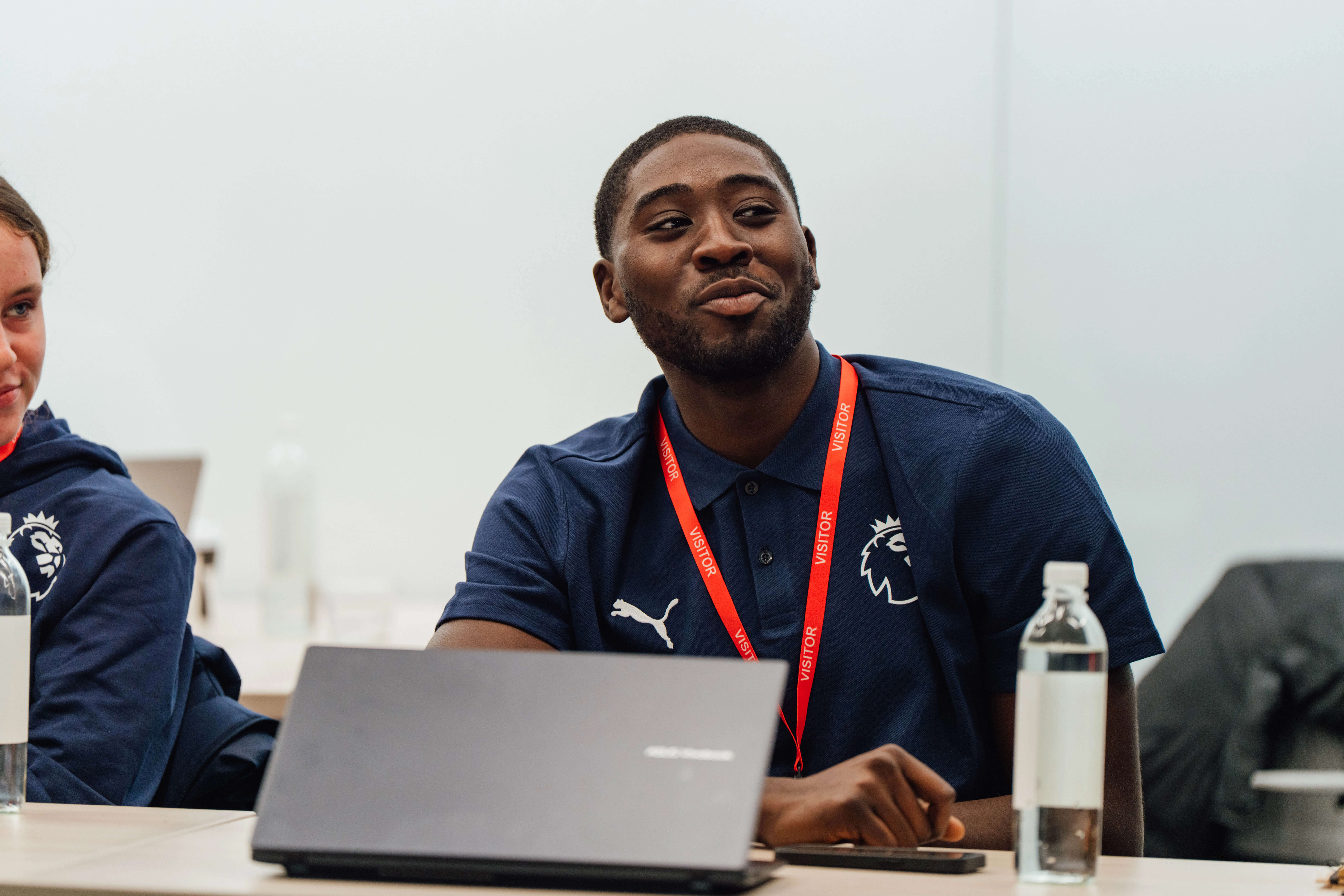Man in a navy blue polo with a Premier League logo and red visitor lanyard sitting at a table with a laptop and bottled water.