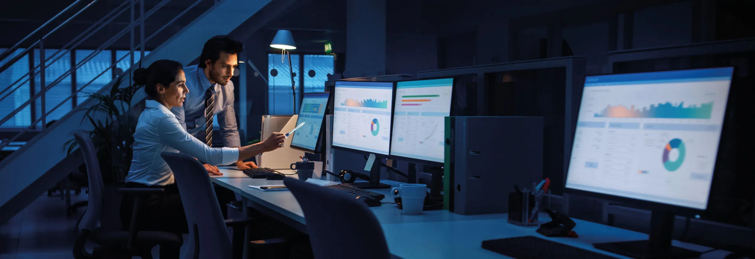 Two logistics professionals working at a bank of dashboard monitors in a control room at night