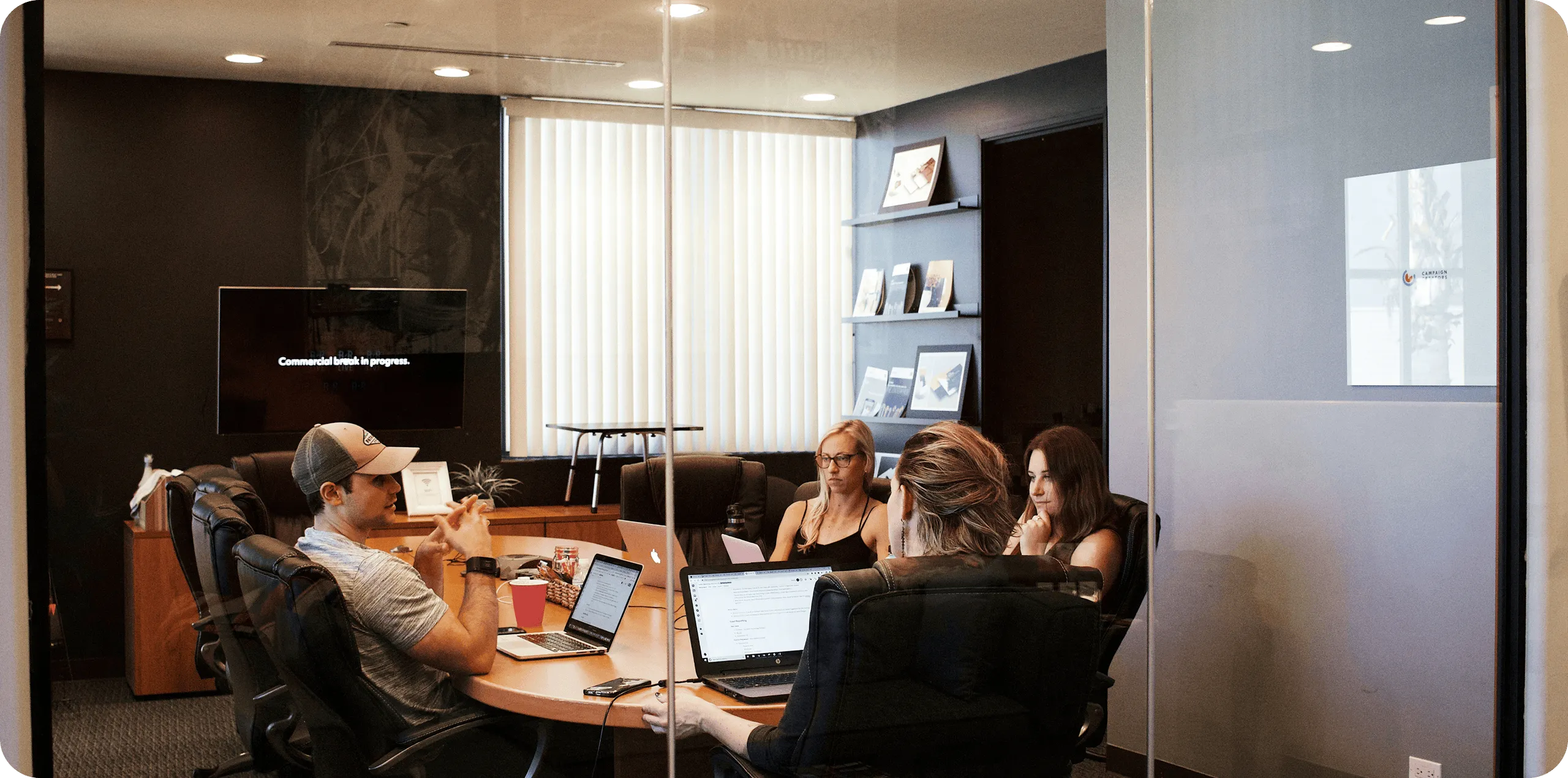 Four people in a glass-walled conference room having a meeting around a round table with laptops and a TV.