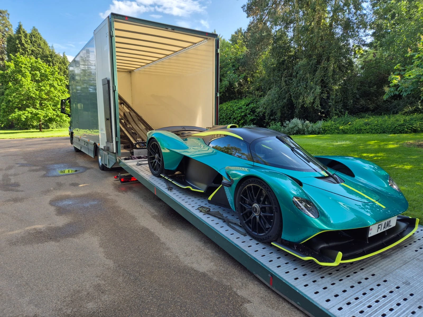 Teal and black sports car with yellow accents loaded on a trailer attached to a green truck in a park setting.