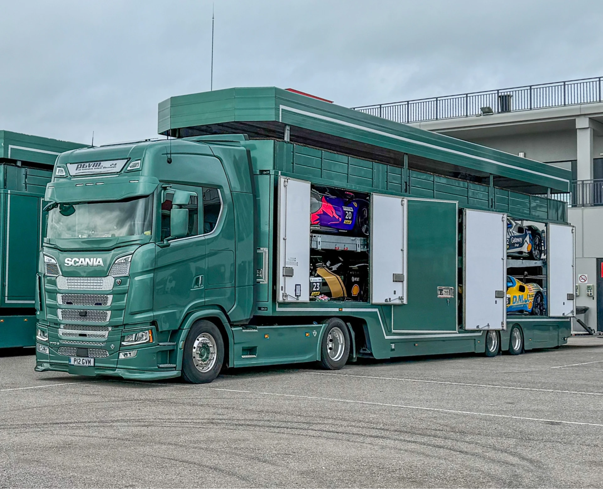Green Scania transport truck with open side doors revealing four colorful racing cars stacked inside at a parking area.