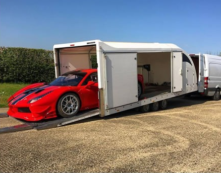 Red sports car with black stripes loaded inside a white enclosed car transporter trailer hitched to a white van.