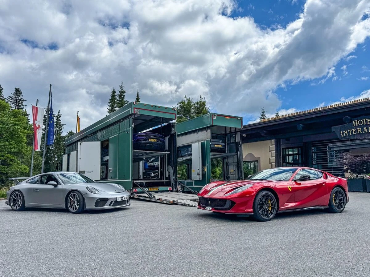 Silver Porsche sports car and red Ferrari parked near green car transport trailers outside a hotel with flags and trees nearby.