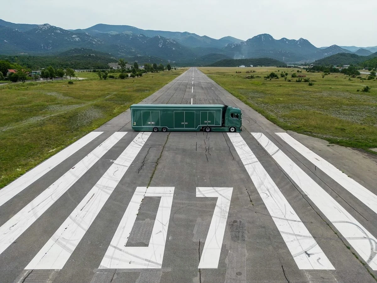 Green semi-truck crossing a runway numbered 107, surrounded by grass fields and mountains in the background.