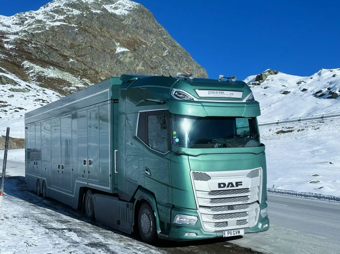 Green DAF semi-truck parked on a snowy mountain road under a clear blue sky.