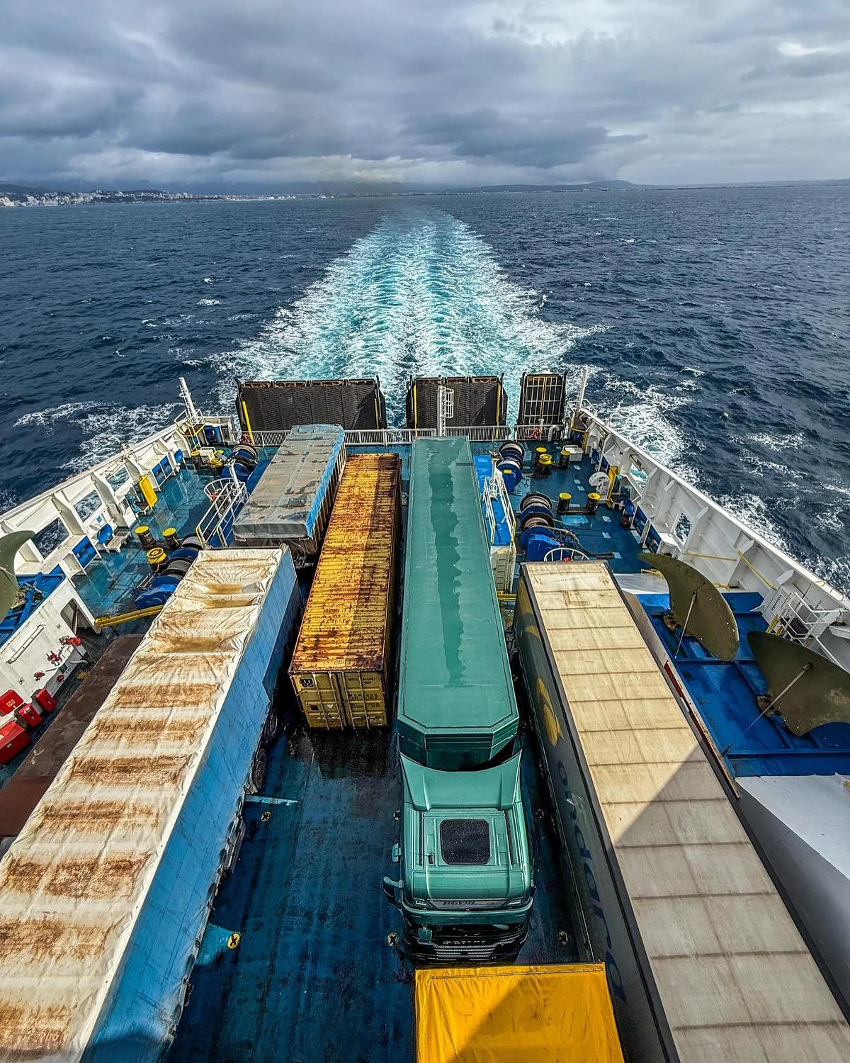 View from a ferry's deck showing several parked trucks and containers with the sea and a cloudy sky in the background.