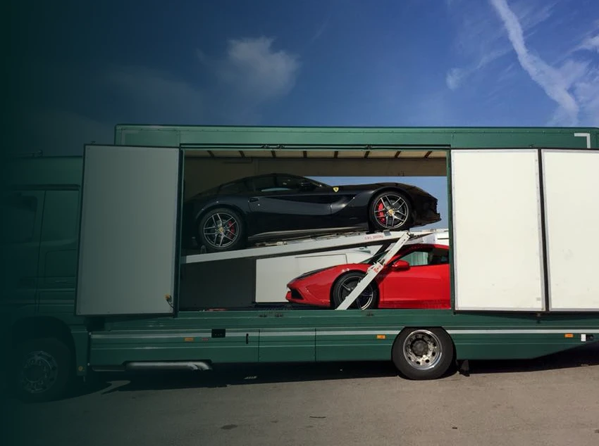 Green truck carrying a red and a black sports car inside a side-open cargo area under a blue sky.