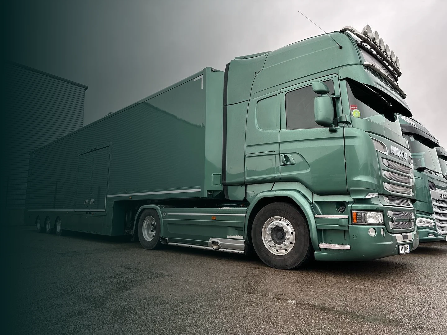 Green semi-truck parked on wet pavement under gray cloudy sky.
