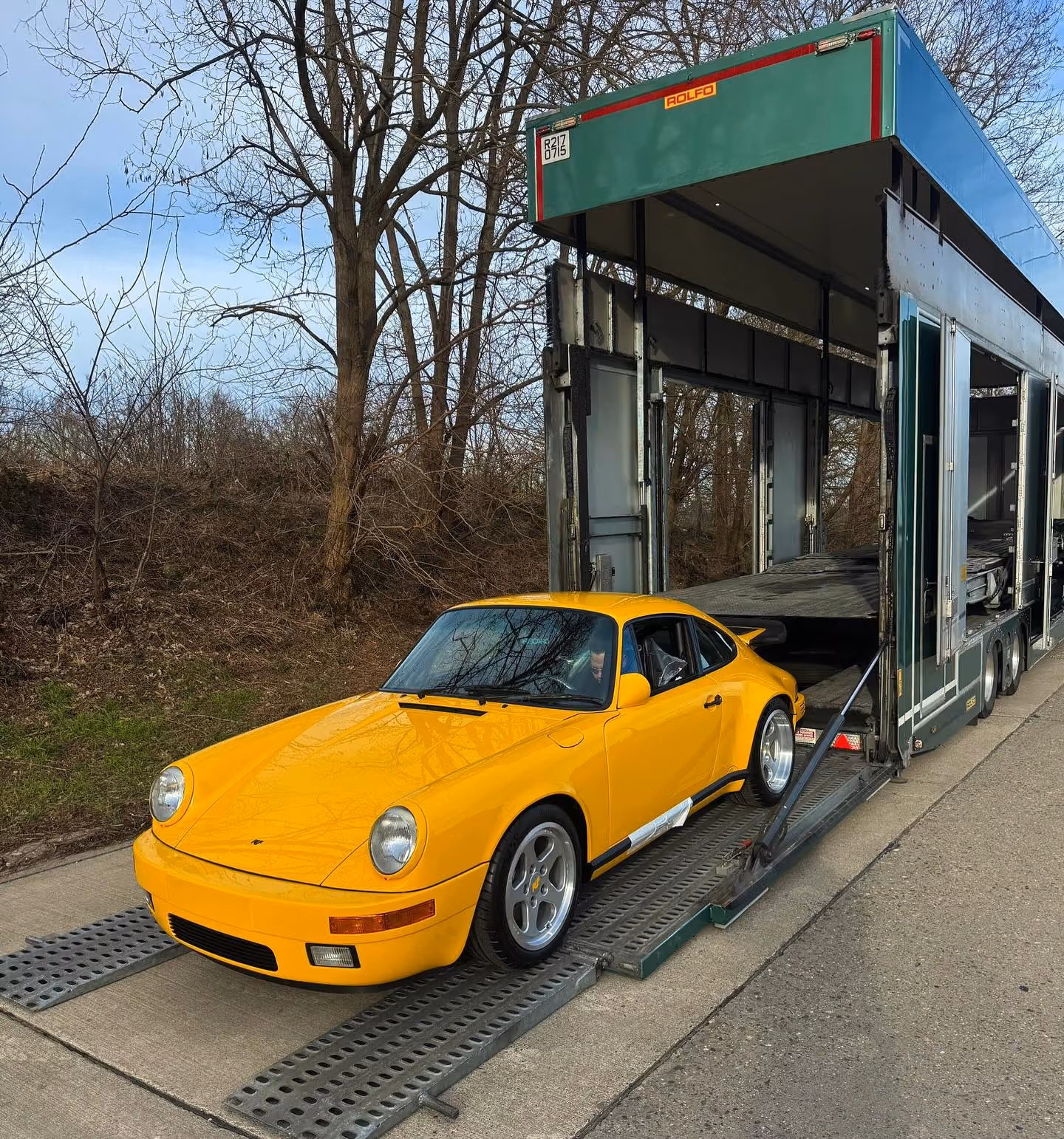 Yellow classic sports car being loaded into a large green enclosed car transport trailer on a concrete surface.