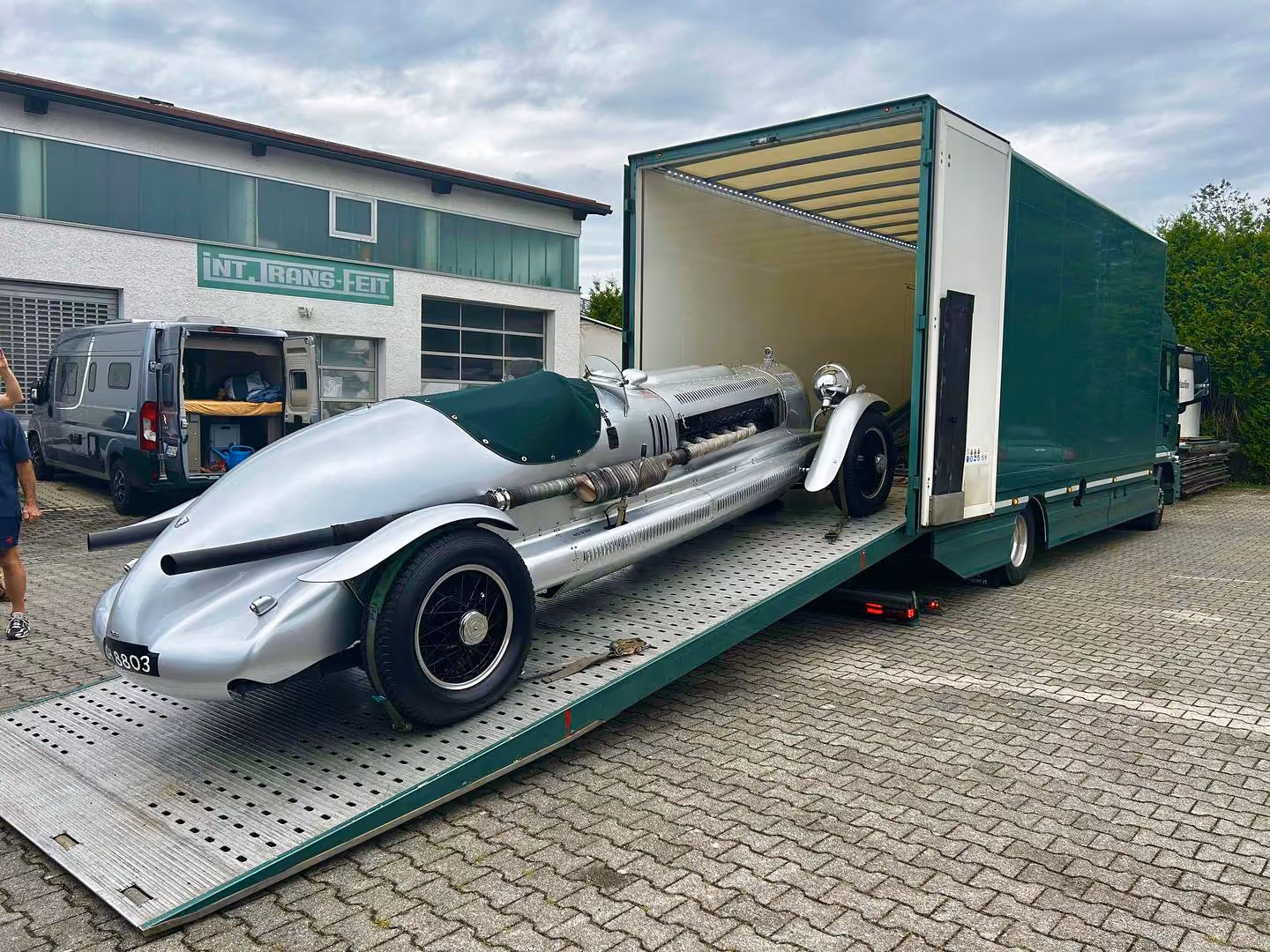 Vintage silver race car loaded on a green enclosed transport truck ramp in front of a building.