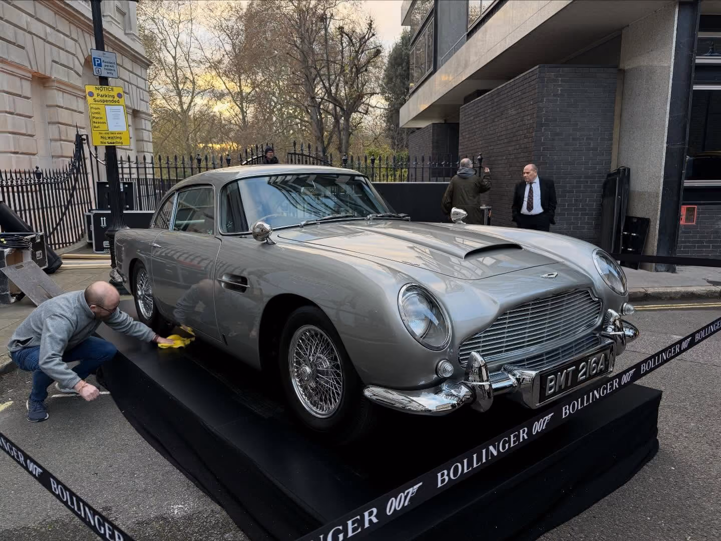 Man polishing a silver classic Aston Martin car with license plate BMT 216A displayed on a raised platform with Bollinger 007 branding.