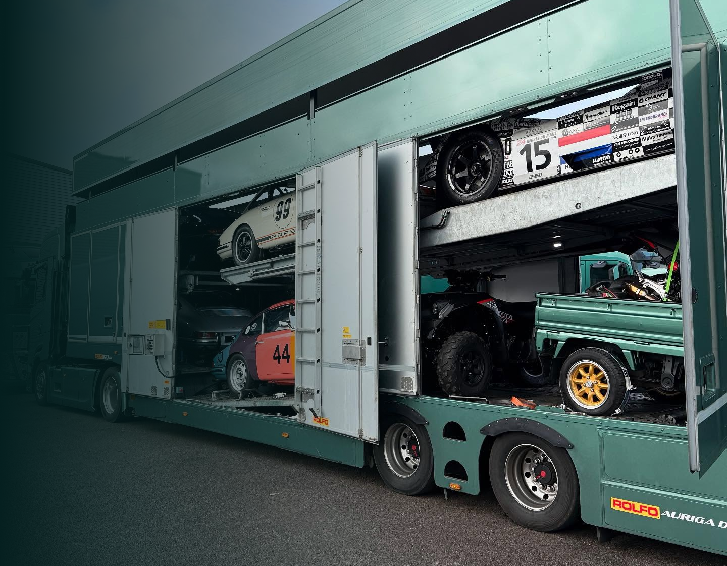 Green truck trailer carrying race cars and an ATV with open side doors showing the vehicles inside.